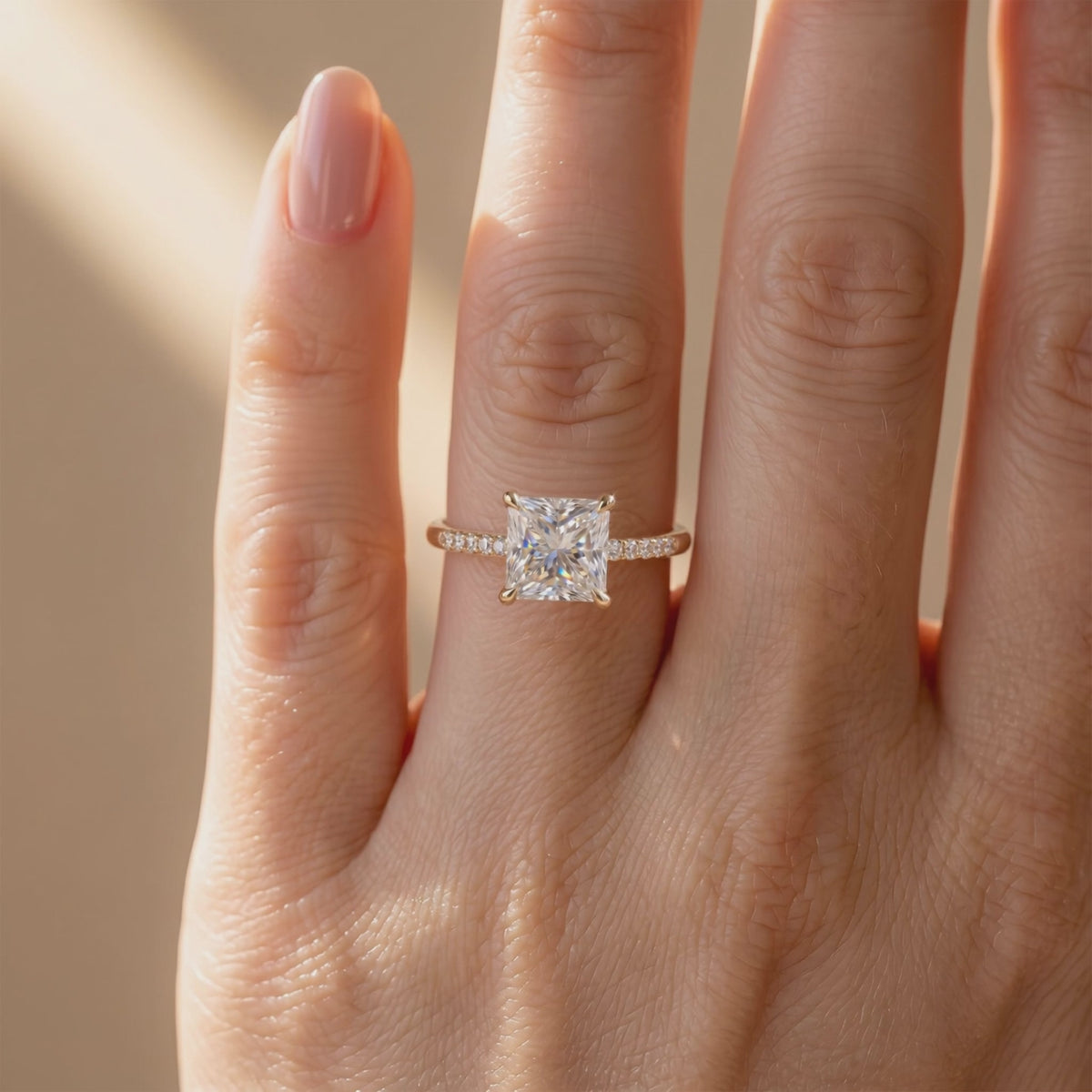 Princess-cut moissanite solitaire ring with a pavé band worn on a person's finger.