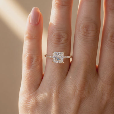 Princess-cut moissanite solitaire ring with a pavé band worn on a person's finger.