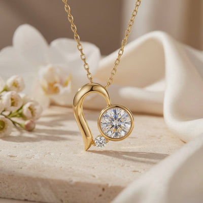 Gold-tone abstract heart pendant necklace with large and small round moissanite stones displayed on a pale stone surface.
