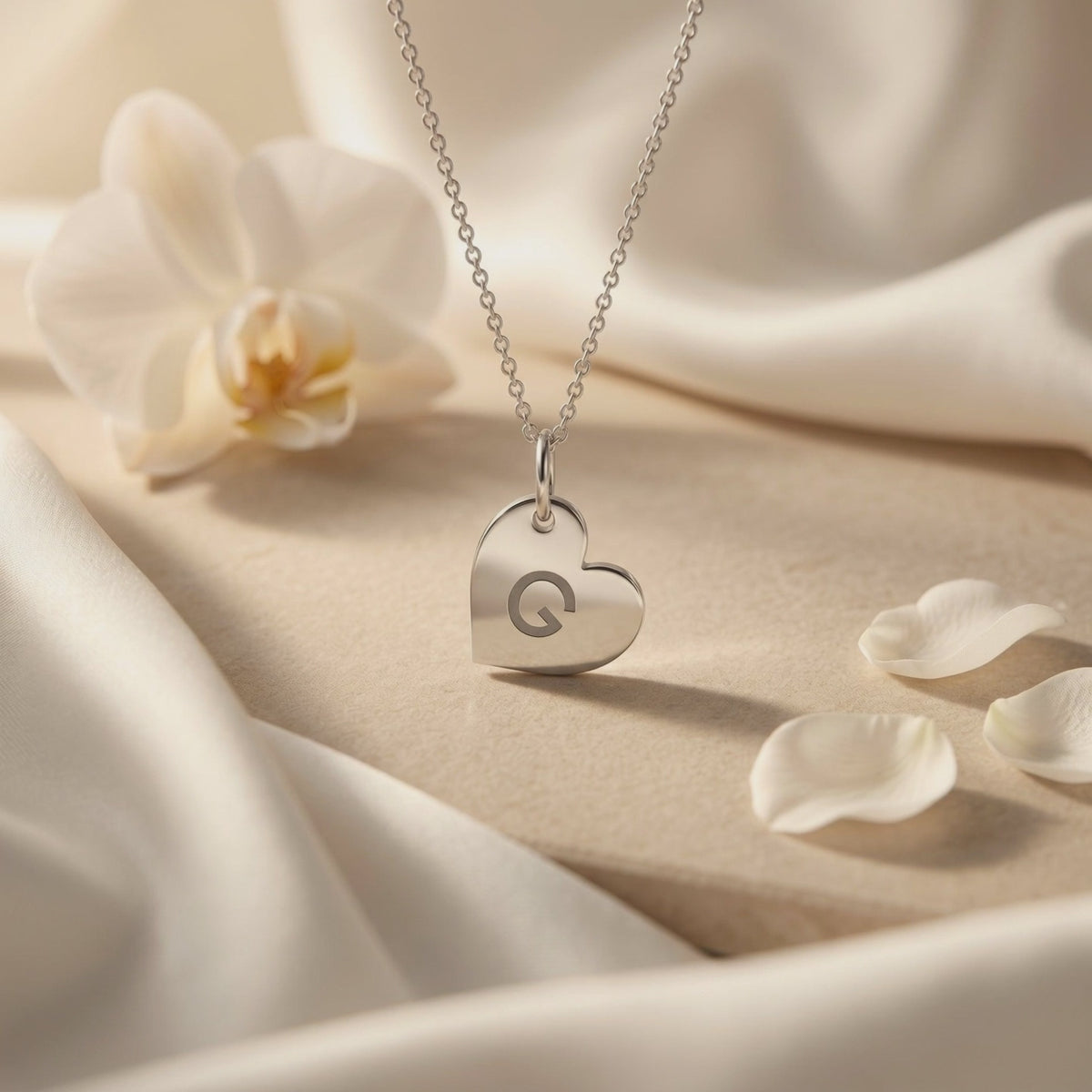 Silver-toned heart pendant engraved with the letter G, displayed on white fabric beside a flower and petals.