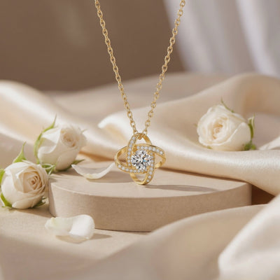 Gold-toned interlocking knot necklace with a clear center stone, resting on a beige pedestal among white roses.