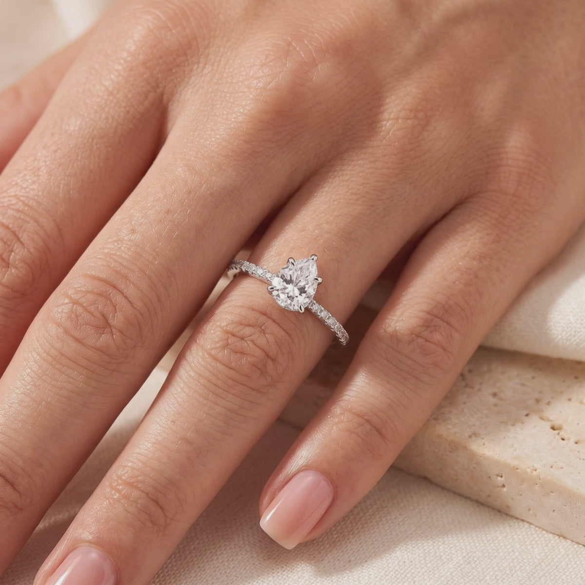 Pear-cut clear stone ring with a pavé band, shown worn on a hand.