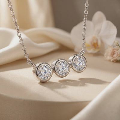 Close-up of a silver necklace with three round bezel-set moissanite stones resting on cream fabric beside flowers.