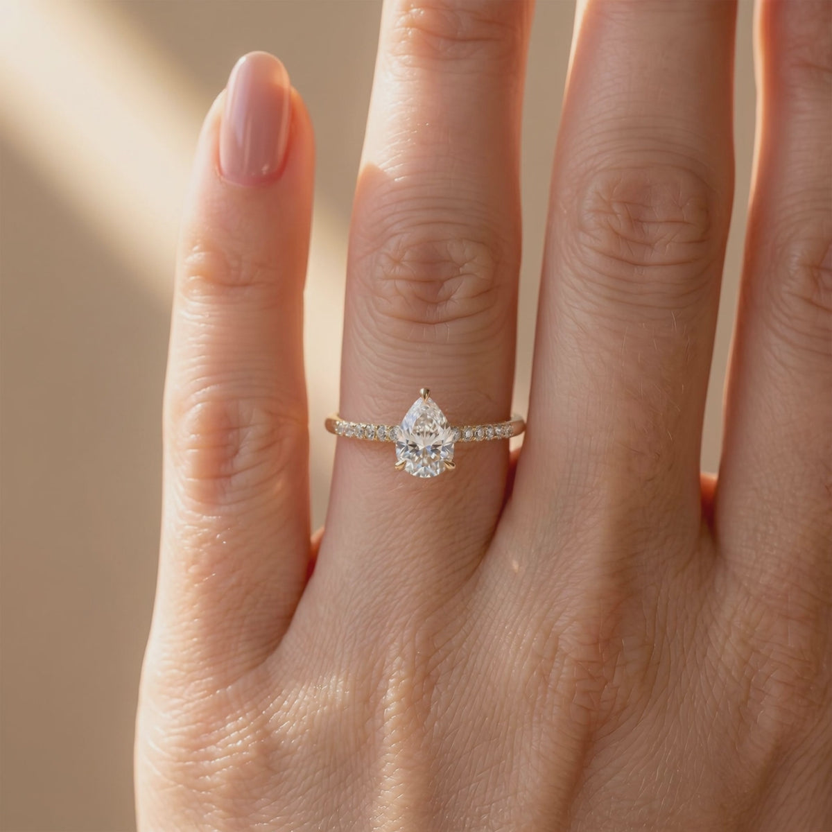 Gold-tone pear-cut solitaire ring with a pavé band worn on a finger in warm light.