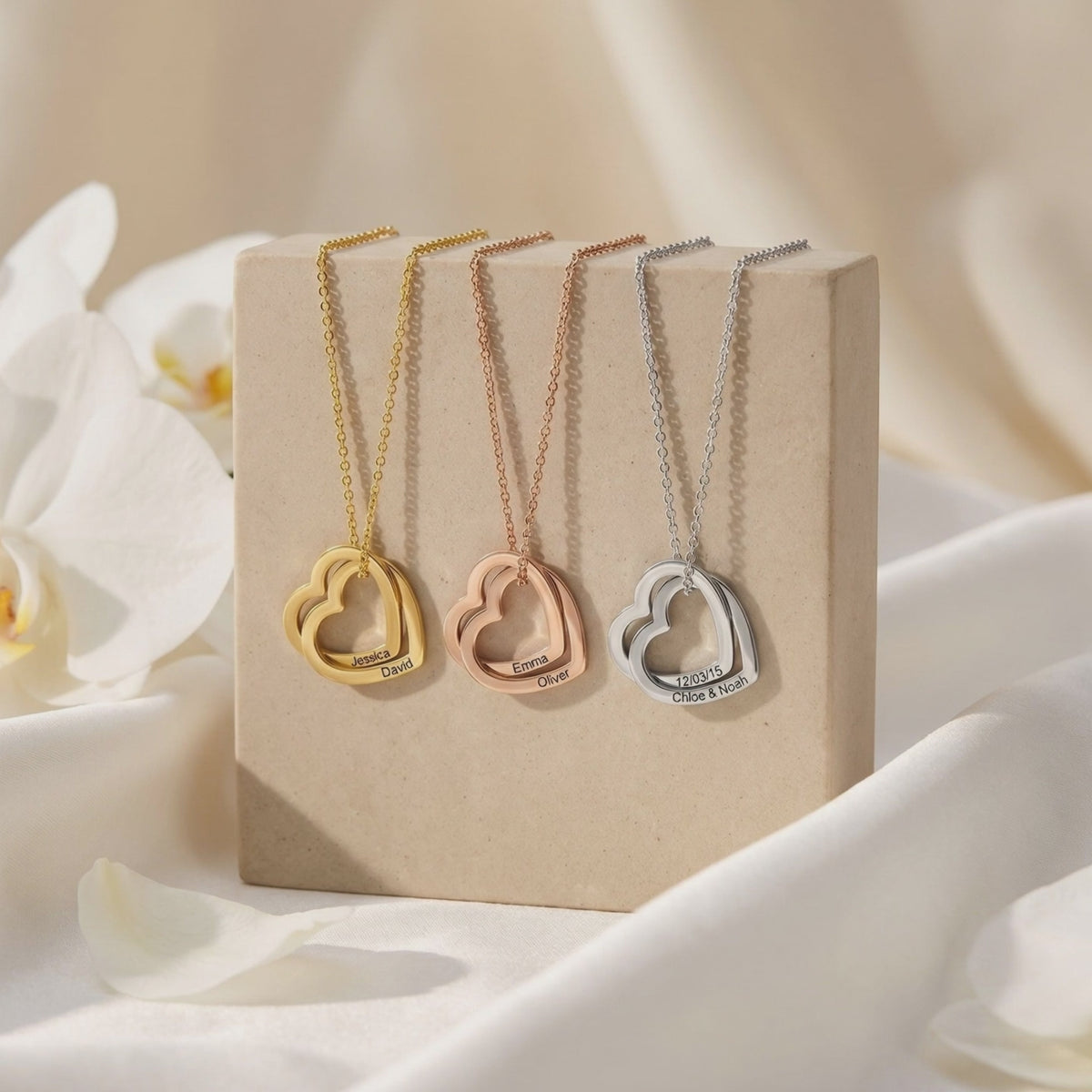 Three engraved double-heart necklaces in gold, rose gold, and silver tones displayed on a neutral stand.
