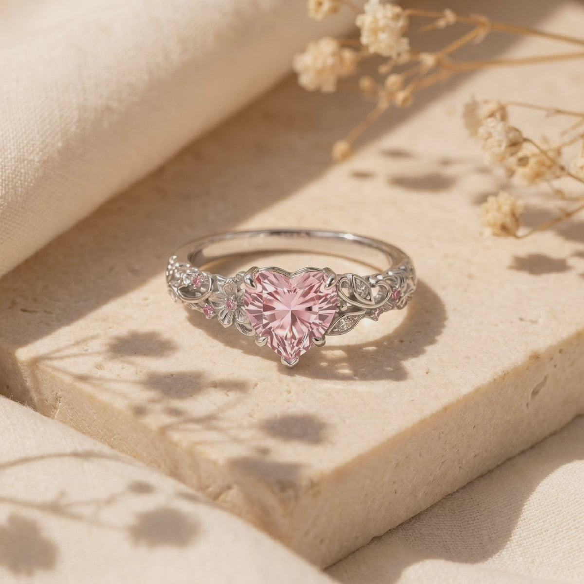 Close-up of a silver ring with a heart-shaped pink centerpiece and floral filigree band resting on a stone surface.