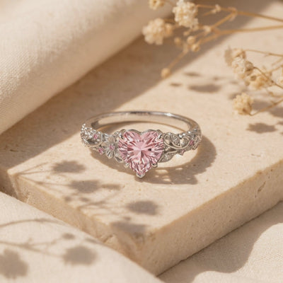 Close-up of a silver ring with a heart-shaped pink centerpiece and floral filigree band resting on a stone surface.