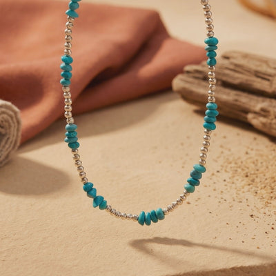 Turquoise and silver-bead necklace hanging above a beige surface, with terracotta fabric and wood in the background.
