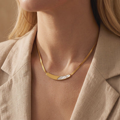 Gold snake chain necklace with a curved mother-of-pearl bar, shown worn on a model in a beige blazer.