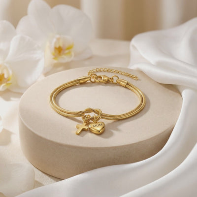 Gold-tone double snake-chain bracelet on a round pedestal, showing clasp, extension chain, cross, and MUM heart charms.