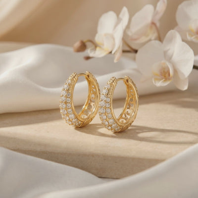 Pair of gold-tone hoop earrings with rows of round moissanite stones displayed beside white orchids.