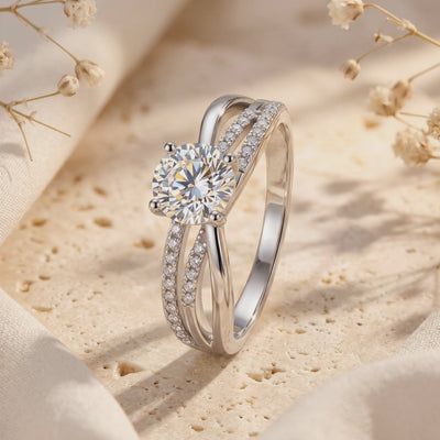 Close-up of a silver crossover ring with a round center stone and pavé bands on a beige stone backdrop.