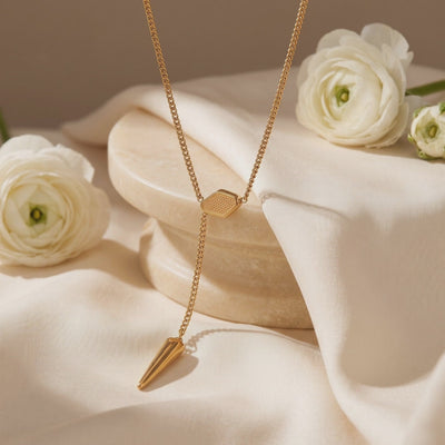 Gold-tone lariat necklace with a textured hexagonal slider and spike pendant, displayed on cream fabric.