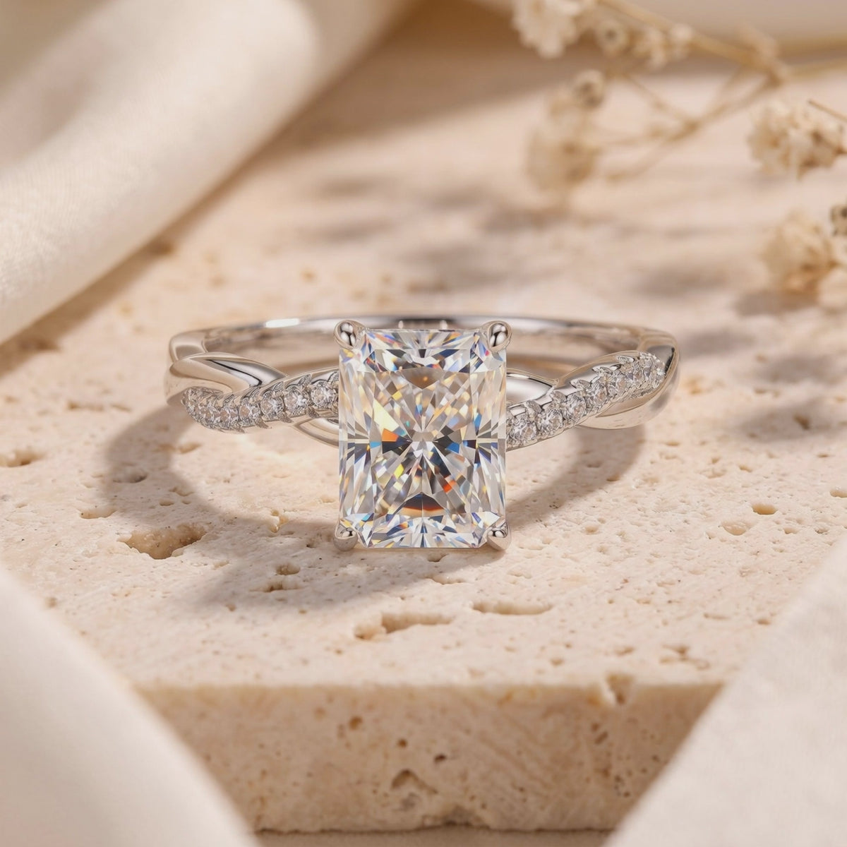 Close-up view of a silver-colored ring with a radiant-cut center stone and twisted pavé band on a textured surface.