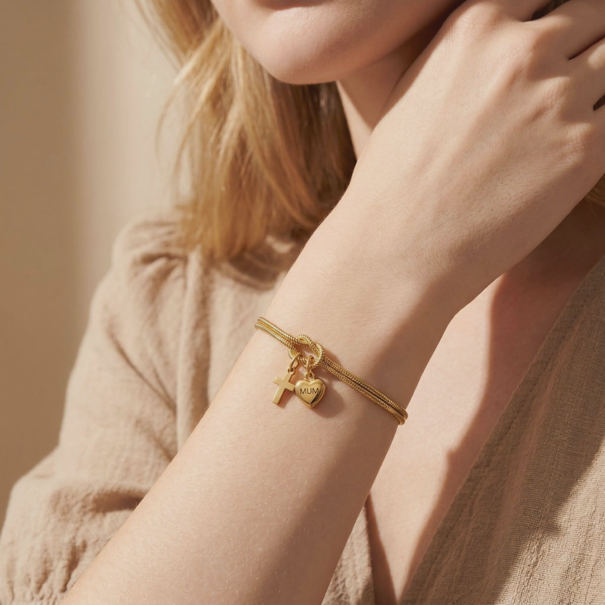 Gold-tone double snake-chain bracelet worn on a wrist, with a knotted center and cross and MUM heart charms.