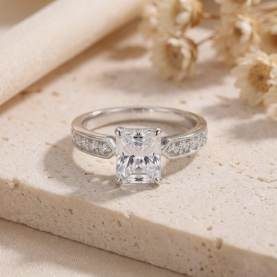 Silver ring with a radiant-cut clear center stone and embellished shoulders, standing upright on a beige stone surface.