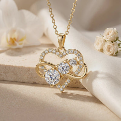 Close-up of a gold-tone heart and infinity moissanite pendant necklace displayed with white flowers.