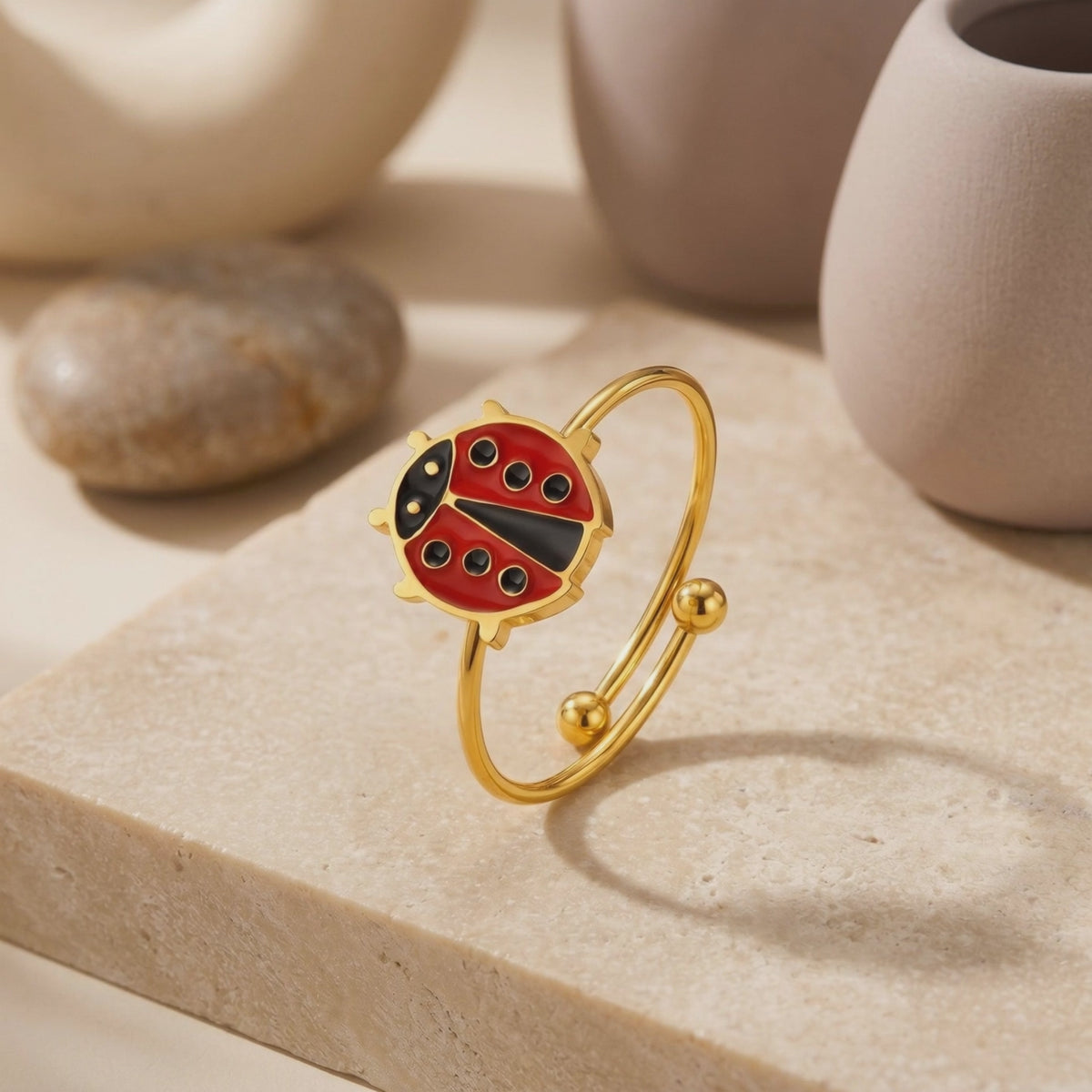 Gold-tone ladybug bypass ring with red and black enamel details, standing on a beige stone surface.