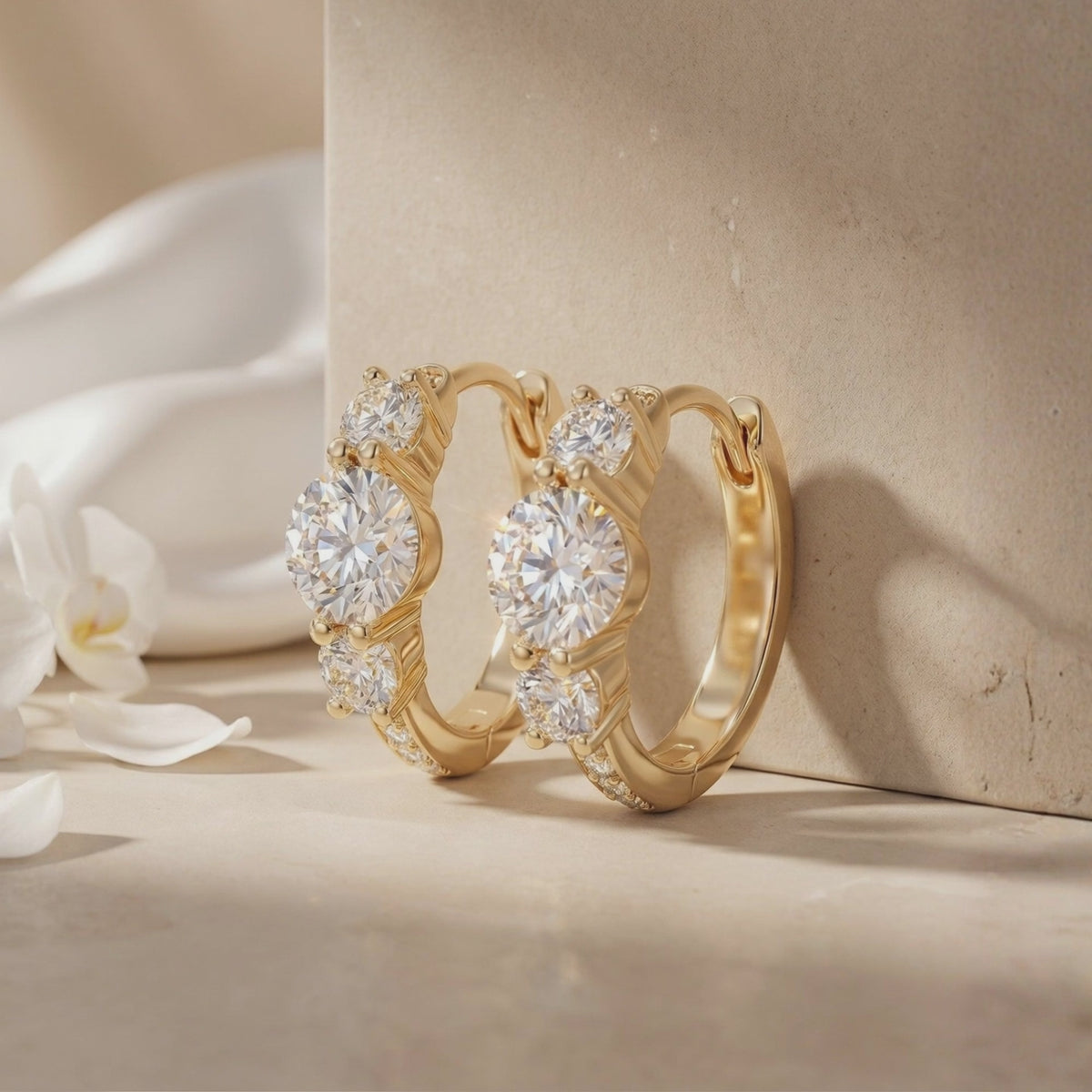 Pair of gold-tone three-stone hoop earrings displayed against a neutral stone block with white petals.