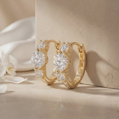 Pair of gold-tone three-stone hoop earrings displayed against a neutral stone block with white petals.