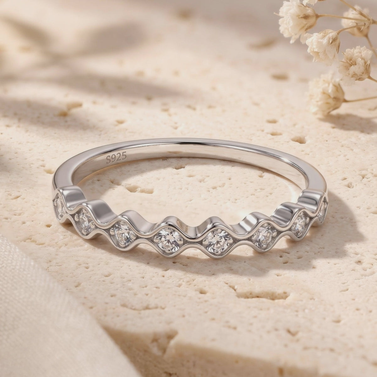Silver wavy ring with round clear stones displayed on a textured cream surface, with an S925 stamp visible inside the band.
