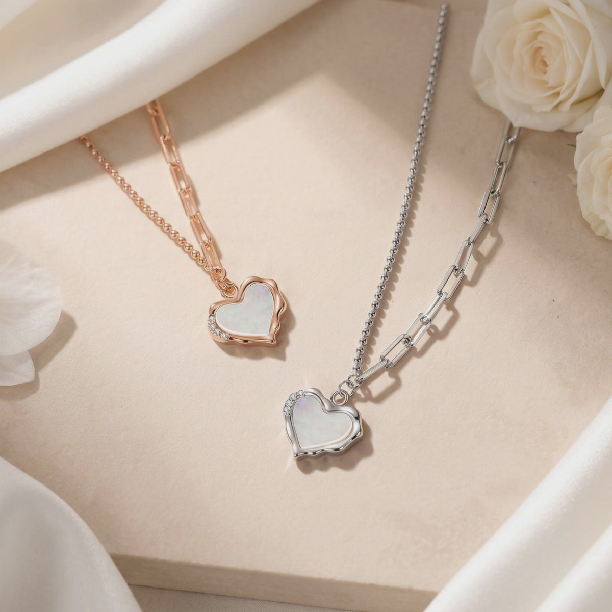 Rose-gold and silver heart pendant necklaces with white opal centers displayed on fabric beside white roses.