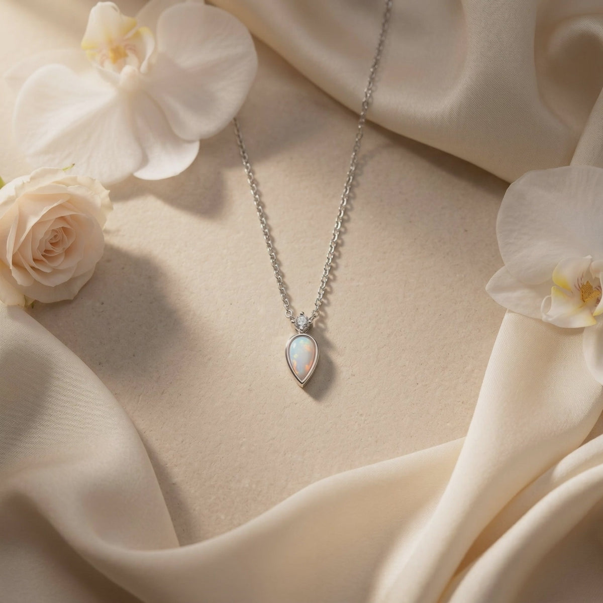 Silver-tone teardrop opal necklace displayed on cream fabric with white flowers.