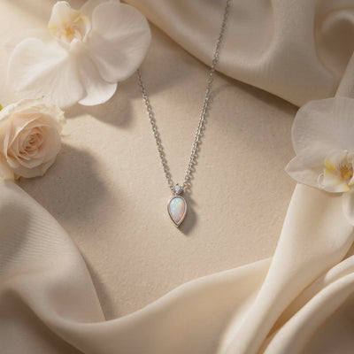 Silver-tone teardrop opal necklace displayed on cream fabric with white flowers.