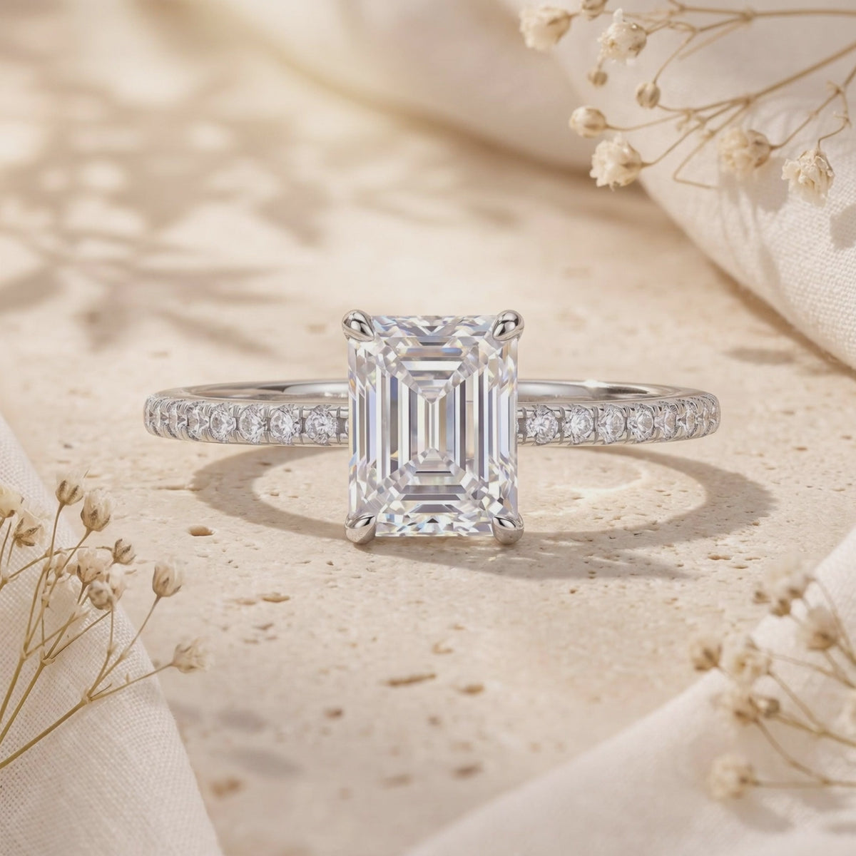 Silver ring with an emerald-cut center stone and pavé band displayed on a beige stone surface.