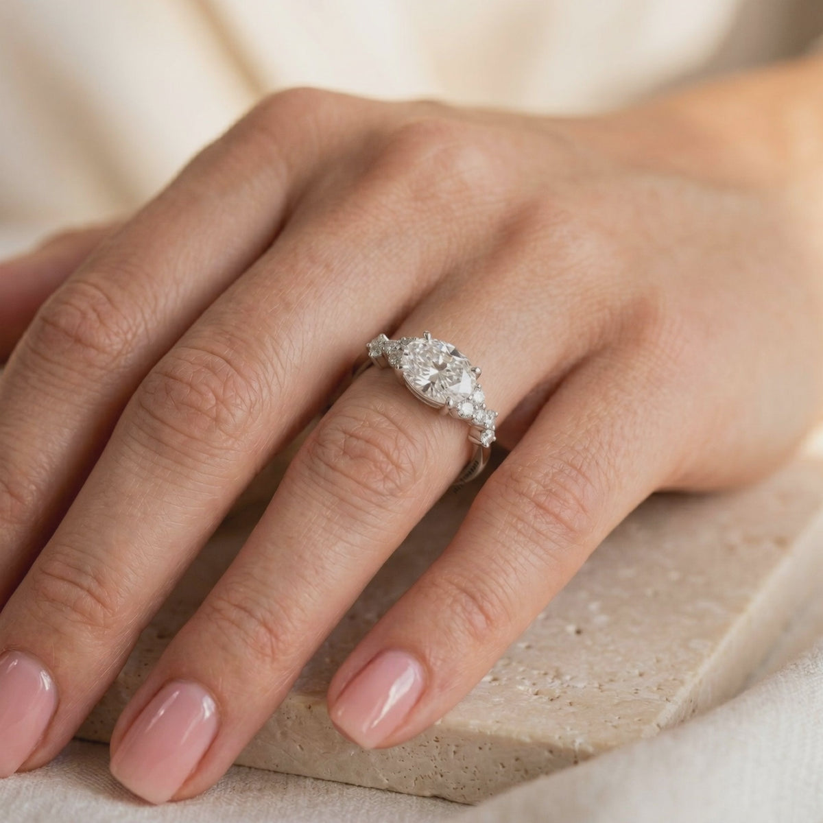 Silver ring with an oval center stone and clustered side accents, worn on a hand with glossy pink nails.