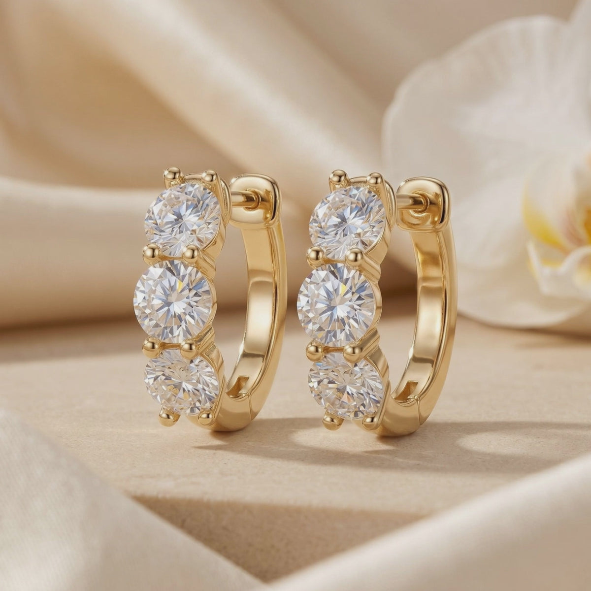 Pair of gold-toned huggie hoop earrings, each with three round prong-set moissanite stones, on a neutral backdrop.