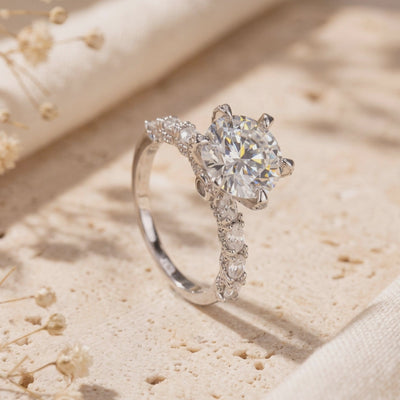 Silver-colored ring standing at an angle on a beige textured surface, with a round clear center stone and accent-set band.