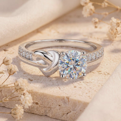 Close-up of a silver love-knot ring with a round clear center stone and pavé shoulders, displayed on a beige textured slab.