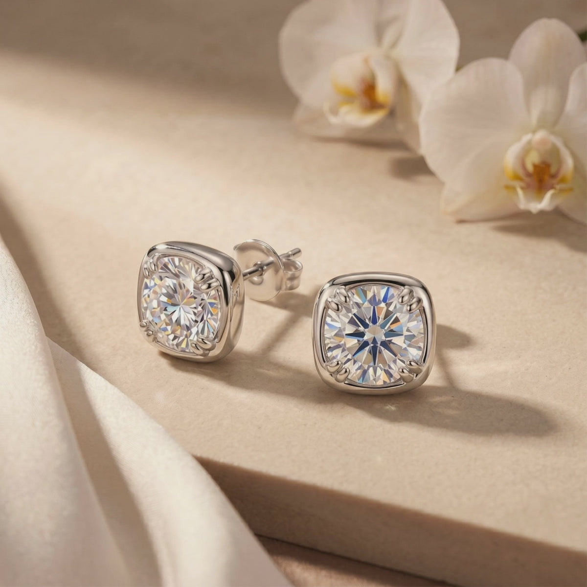 Pair of silver cushion-halo moissanite stud earrings displayed on a light-colored surface beside white orchids.