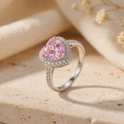 Close-up of a silver ring with a heart-shaped pink center and sparkling halo, standing upright on a textured beige surface.