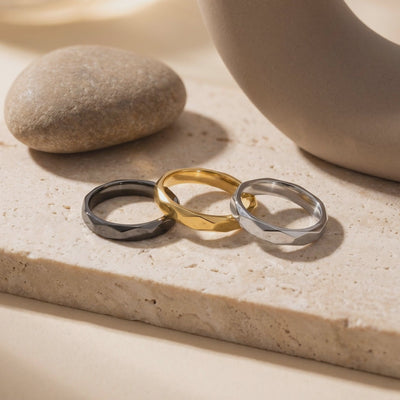 Three faceted band rings in black, gold-tone, and silver-tone on a stone surface.