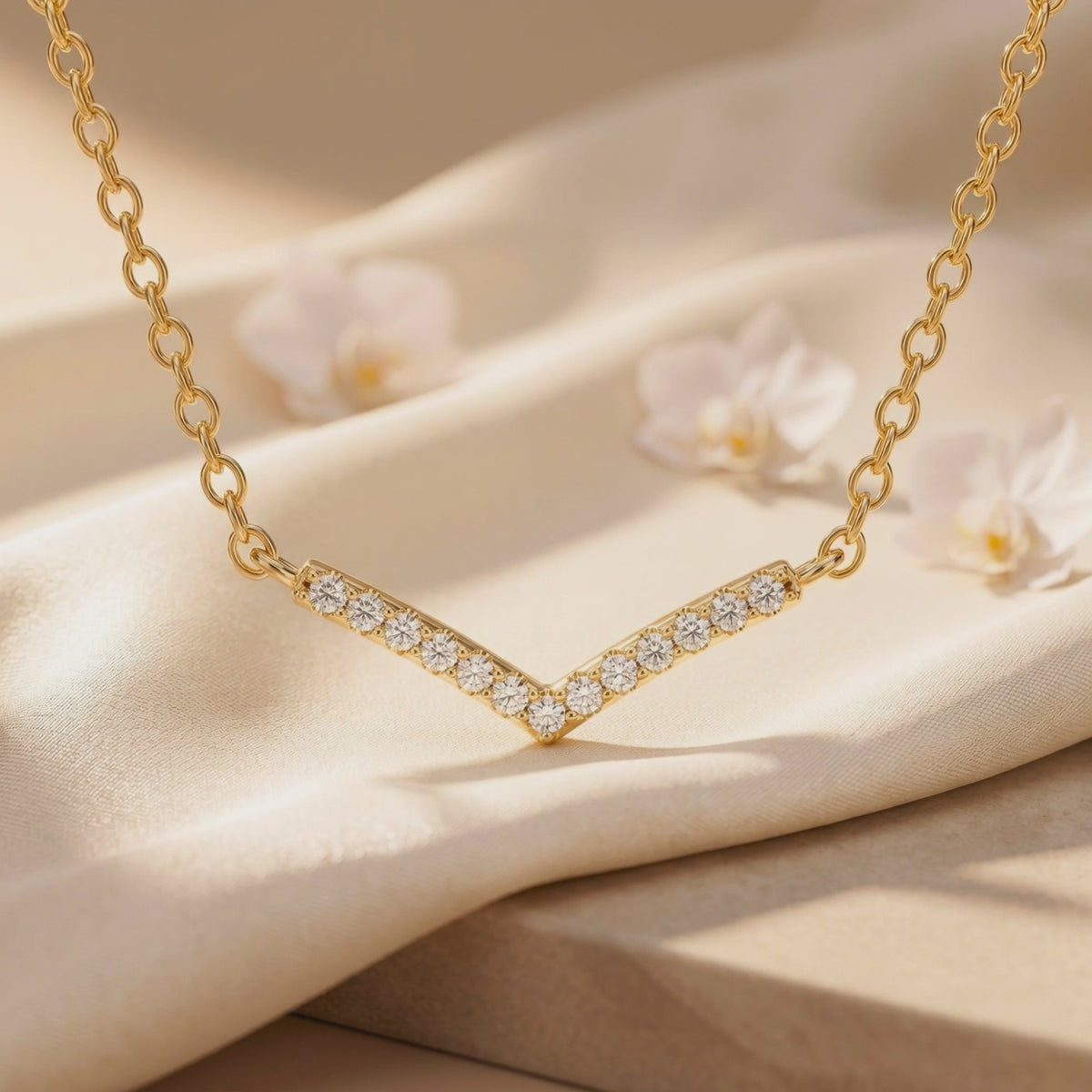 Close-up of a gold-tone chevron pendant with round clear stones on cream fabric, with white flowers in the background.