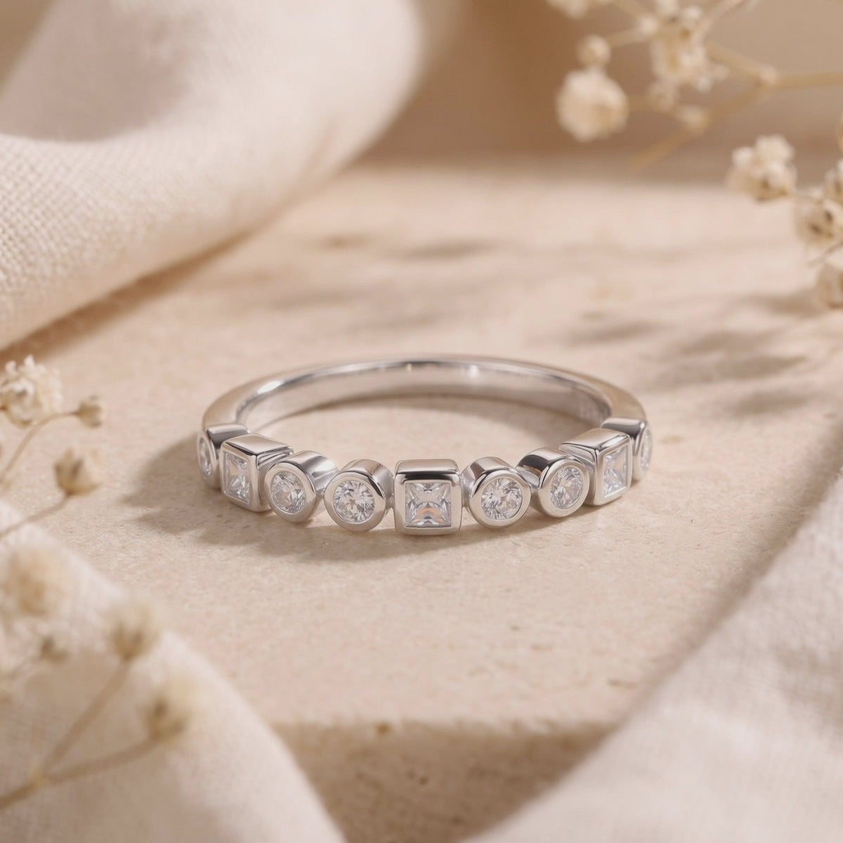 Silver ring with alternating round and square bezel-set clear stones displayed on beige fabric.