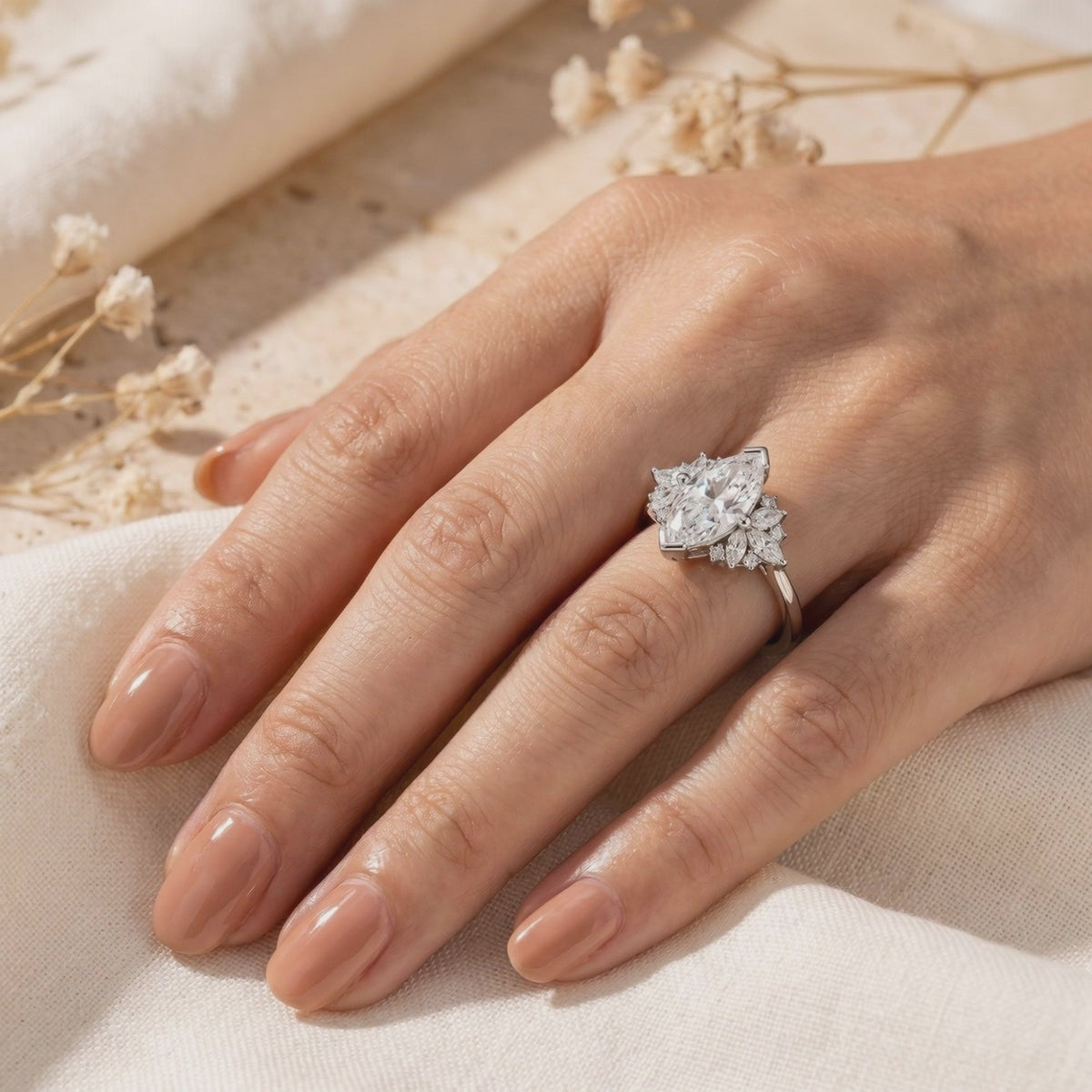 Marquise moissanite cluster ring in silver, worn on a hand over neutral fabric beside dried flowers.