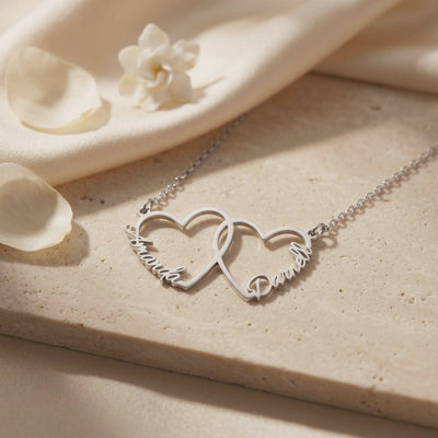 Silver-toned interlocking heart necklace with Amanda and Daniel, laid on a pale textured block with white petals.