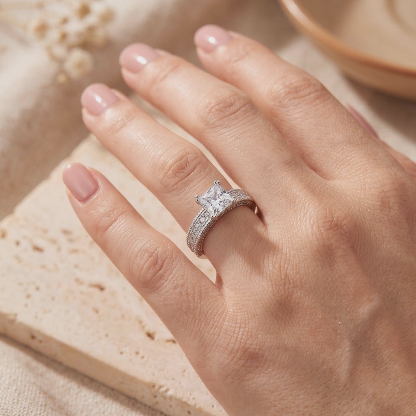 Silver ring with a princess-cut center stone and pavé band worn on a hand over a textured surface.