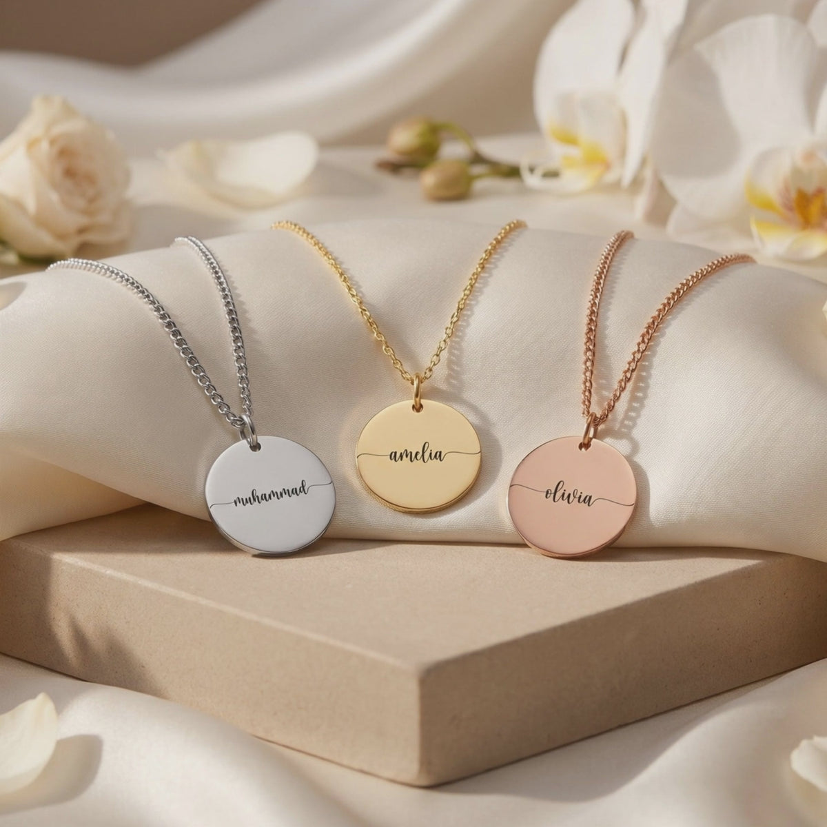 Three round Eloise disc pendant necklaces in silver, gold, and rose gold, engraved “muhammad,” “amelia,” and “olivia.”