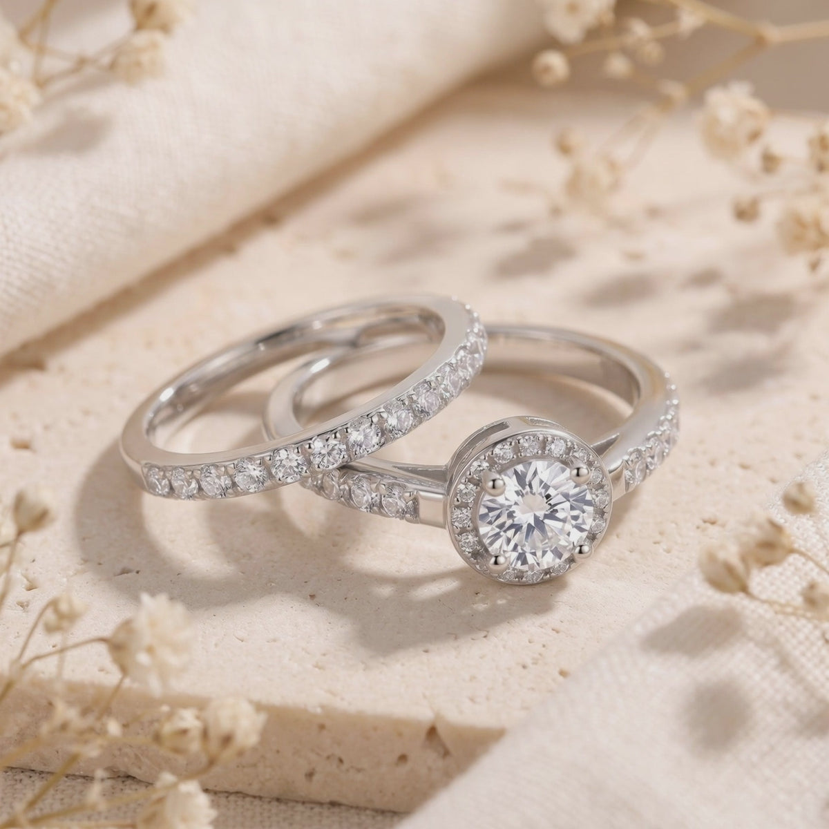 Silver halo ring set with a round center stone and pavé bands displayed on a cream textured surface with dried flowers.