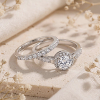 Silver halo ring set with a round center stone and pavé bands displayed on a cream textured surface with dried flowers.