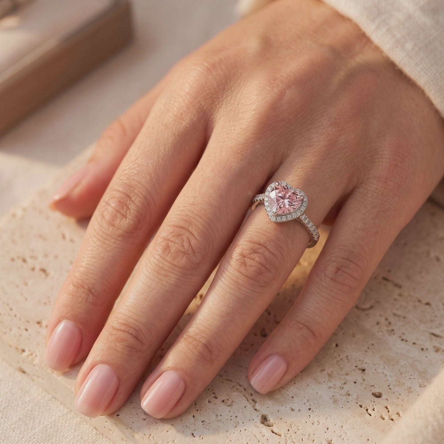 Silver ring with a heart-shaped pink center and sparkling halo, worn on a hand over a textured beige surface.